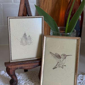 May include: Two framed art prints with gold-colored frames. One features a detailed illustration of a hummingbird in flight, and the other depicts two stylized pine trees. The frames are displayed on a small, ornate wooden easel.