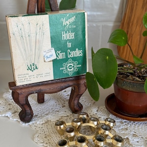 May include: A vintage candle holder set with a box that reads "Vogue 12 Cup Holder for Slim Candles". The box is on a small wooden easel. A gold-colored metal candle holder is in the foreground. The box has a green and white design.