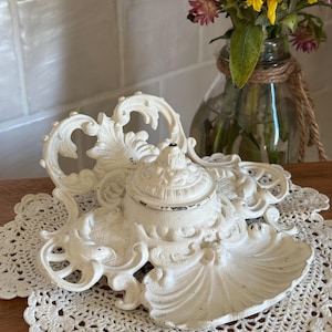 May include: Off-white, ornate inkwell with a lid, set on a cream-colored doily. The inkwell features a decorative, scroll-like design. A glass vase with sunflowers and pink flowers is in the background.