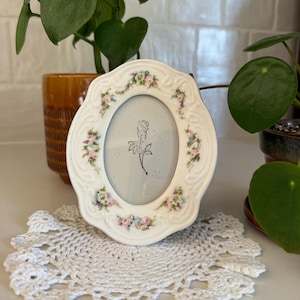 May include: An oval-shaped white ceramic picture frame with a scalloped edge and embossed scrollwork details. The frame is decorated with small, hand-painted floral clusters in pink, blue, and green. A black line drawing of a single rose is inside.