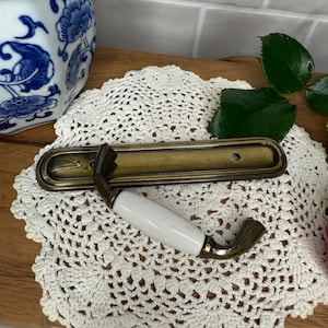 May include: Antique brass cabinet pull with a white ceramic handle. The pull has a rectangular backplate and a curved handle. The hardware is likely for a drawer or cabinet door.