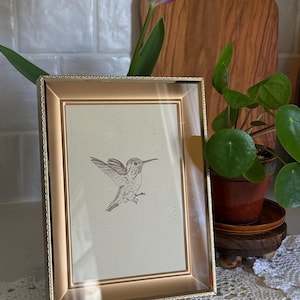 May include: A gold-colored rectangular picture frame with a hummingbird drawing inside. A purple tulip and green plant are in the background. The frame is on a white surface with a lace doily.