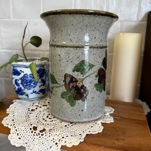 May include: A speckled ceramic vase with a wide opening and a floral design in brown, green, and burgundy. The vase is on a wooden surface with a white lace doily. A small potted plant and a candle are in the background.