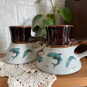 May include: Two ceramic mugs with a shark theme. The mugs have a brown top and a white base with teal shark illustrations. The handles are brown and the mugs are sitting on a white lace doily.