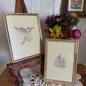 May include: Two framed prints with gold-colored frames. One features a hummingbird illustration, the other a pair of trees. The frames are displayed on a wooden easel and a table with a lace doily. A small framed picture and a bouquet of flowers are in the background.