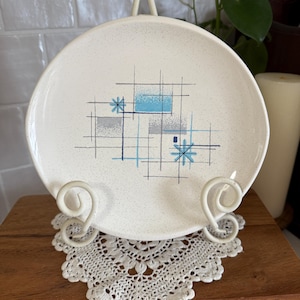 May include: A white ceramic plate with a speckled surface, featuring a mid-century modern design in blue, gray, and black geometric shapes. The plate is displayed on a white metal stand and a lace doily.