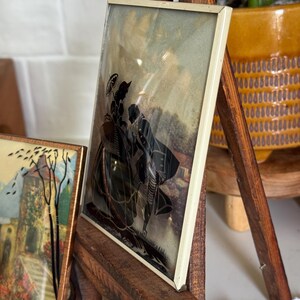 May include: A framed silhouette art piece featuring a black depiction of two figures with an umbrella. The artwork is set against a muted background and framed in a cream-colored border. The frame is displayed on a wooden easel.