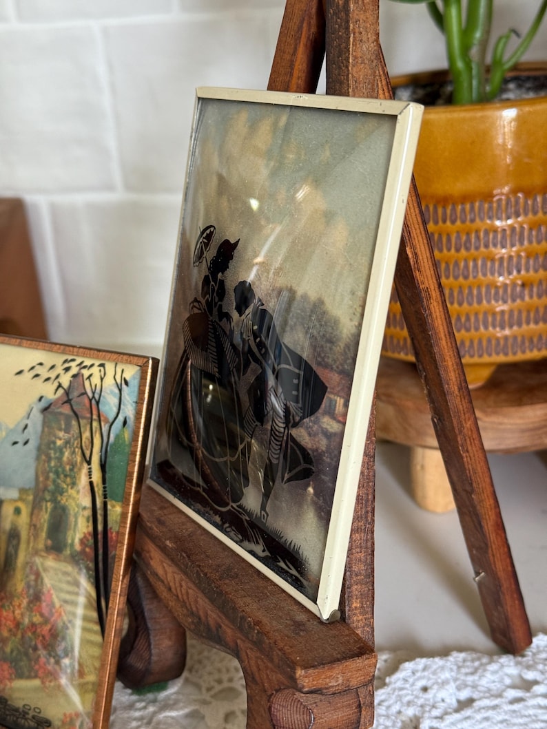 May include: A framed silhouette art piece featuring a black depiction of two figures with an umbrella. The artwork is set against a muted background and framed in a cream-colored border. The frame is displayed on a wooden easel.