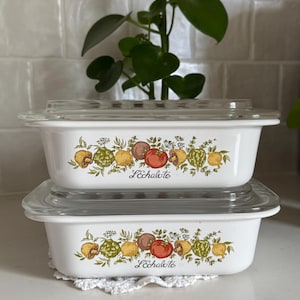 May include: Two white rectangular casserole dishes with clear glass lids are stacked. Each dish features a decorative band of colorful vegetables and the text "L'Echalote". The dishes are on a white surface.
