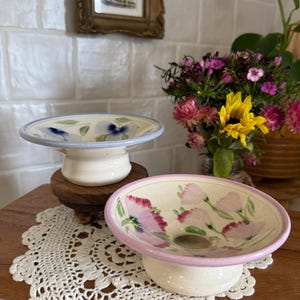 May include: Two ceramic pedestal bowls. One bowl has a light blue rim and blue floral design. The other bowl has a pink rim and pink floral design. Both bowls are white with a pedestal base. The bowls are on a wooden surface.