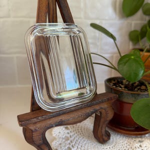 May include: A rectangular clear glass tray featuring a series of vertical ribbed lines along its center. The tray has rounded corners and a smooth, raised outer rim, showcasing a transparent design that reflects light across its textured surface.