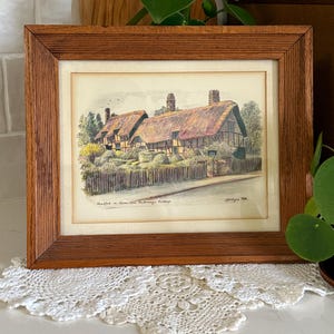 May include: A framed print of a thatched-roof cottage with a wooden fence. The artwork is in a brown wooden frame. The print features a detailed illustration with muted colors, depicting a quaint rural scene. The frame is made of wood.