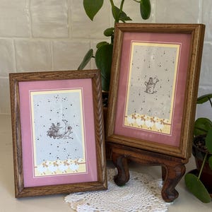 May include: Two rectangular wooden frames with dark brown grain, each containing a light gray print with a pink mat. The left print shows a goose with a cart of flowers, while the right print features a watering can. Both have a row of white bows.