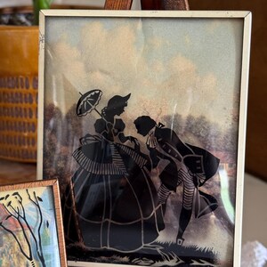 May include: A framed silhouette art piece depicting a man bowing to a woman holding an umbrella. The figures are black against a backdrop of a cloudy sky and trees. The frame is a thin, off-white metal.