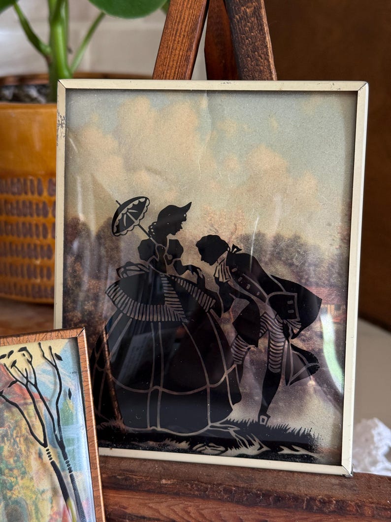 May include: A framed silhouette art piece depicting a man bowing to a woman holding an umbrella. The figures are black against a backdrop of a cloudy sky and trees. The frame is a thin, off-white metal.