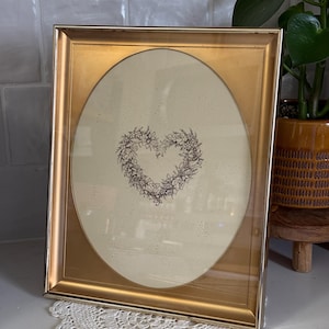 May include: A gold-framed picture with an oval opening. The picture inside features a heart-shaped floral design in brown against a cream background. The frame is rectangular with a gold finish. The picture is displayed on a white surface with a lace doily.