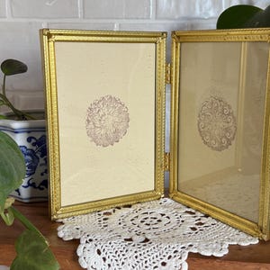 May include: A gold-colored, hinged picture frame with two panels. Each panel holds a cream-colored background with a floral design. The frame is resting on a wooden surface with a white lace doily.