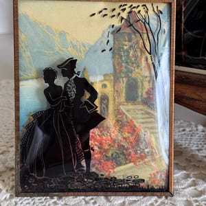 May include: A framed silhouette art piece featuring a couple in period attire. The black silhouettes are set against a backdrop of a landscape with mountains, a lake, and a building. The frame is a warm brown color.