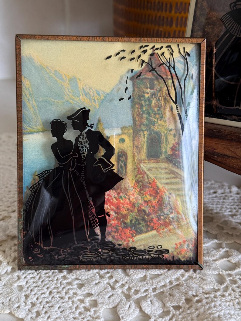 May include: A framed silhouette art piece featuring a couple in period attire. The black silhouettes are set against a backdrop of a landscape with mountains, a lake, and a building. The frame is a warm brown color.
