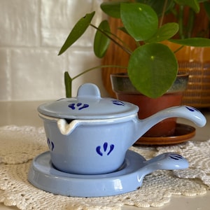 May include: A light blue enamelware sauce pot with a matching lid and a small, attached saucer. The pot and saucer have blue floral accents. The pot has a long handle and a spout. The set is sitting on a white lace doily.