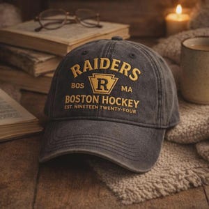 May include: A dark gray baseball cap with the word "RAIDERS" embroidered in gold, along with "BOS MA BOSTON HOCKEY EST. NINETEEN TWENTY-FOUR". The cap has a curved brim and a classic six-panel design.