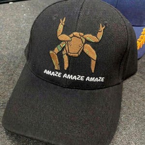 May include: A black baseball cap featuring an embroidered tan, multi-legged creature with green accents on the front panel. Below the creature, the text "AMAZE AMAZE AMAZE" is embroidered in white capital letters. The cap has a structured crown.