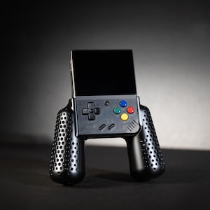 May include: A black handheld gaming console with a screen and buttons. The console has two black controllers attached to the sides.