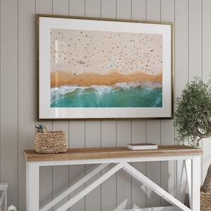 May include: A framed aerial photograph of a sandy beach with numerous people scattered across the shore, meeting the turquoise ocean waves. The print is encased in a thin metallic frame and mounted on a wall with vertical paneling details.