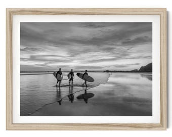 May include: A black and white framed photograph depicting three individuals carrying surfboards across a wet, reflective beach at dusk. The scene features a cloudy sky, calm water, and distant cliffs, all enclosed within a light-toned wooden frame.