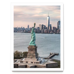 May include: A photographic print featuring the Statue of Liberty in a verdigris copper finish standing on a stone pedestal. The New York City skyline, including One World Trade Center, is visible in the background across the water under a soft sky.