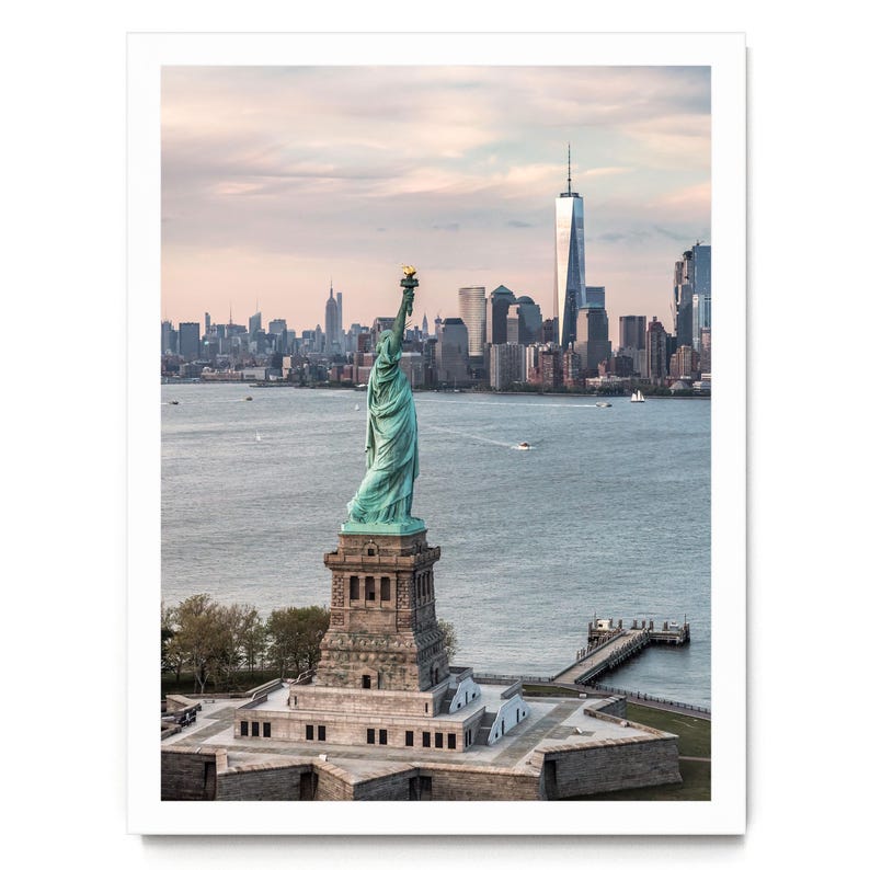 May include: A photographic print featuring the Statue of Liberty in a verdigris copper finish standing on a stone pedestal. The New York City skyline, including One World Trade Center, is visible in the background across the water under a soft sky.