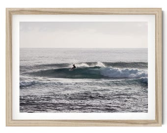 May include: A framed wall art print featuring a photograph of a surfer riding a wave in the ocean. The scene shows a dark silhouette of a person on a surfboard against a vast, light-colored sea and sky, enclosed in a light-toned wooden frame.