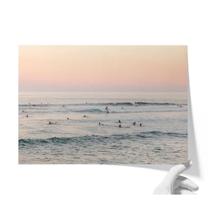 May include: A landscape photograph of a calm ocean at sunset with a soft pink and orange sky. Numerous surfers are scattered across the water, some paddling on boards and others waiting for waves in the gentle, rolling blue-grey sea.