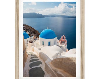 Könnte beinhalten: Ein gerahmter Wandkunstdruck mit einer Landschaft von Santorin, Griechenland, mit weißen Gebäuden, leuchtend blauen Kuppeln und einer Steintreppe, die zum tiefblauen Ägäischen Meer unter einem bewölkten Himmel führt, in einem hellen Holzrahmen.