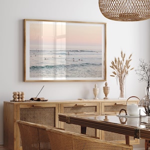 a serene beach scene with a large framed picture hanging on the wall above a wooden cabinet. The cabinet has a glass top and is adorned with various decorative items, including a vase and a bowl. The dining table is positioned in front of the cabinet, creating a cozy and inviting atmosphere in the room.