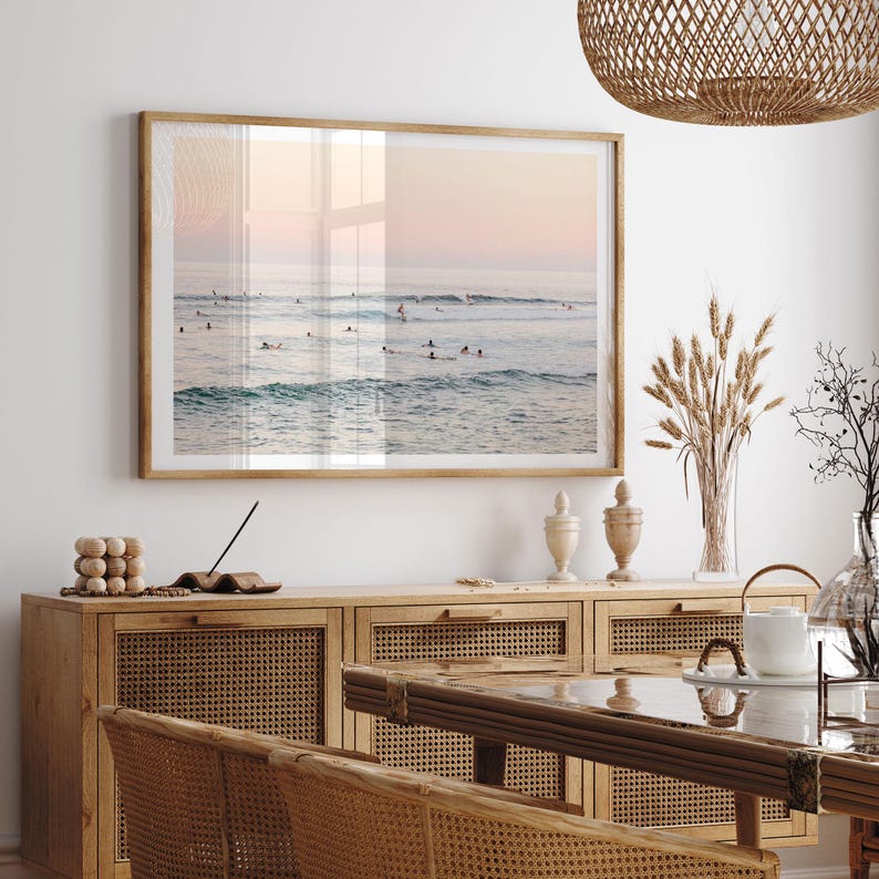 a serene beach scene with a large framed picture hanging on the wall above a wooden cabinet. The cabinet has a glass top and is adorned with various decorative items, including a vase and a bowl. The dining table is positioned in front of the cabinet, creating a cozy and inviting atmosphere in the room.