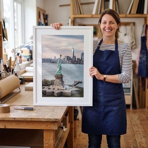 May include: A framed vertical photograph of the Statue of Liberty standing on Liberty Island with the New York City skyline and One World Trade Center visible in the background under a soft, light-filled sky during the daytime.