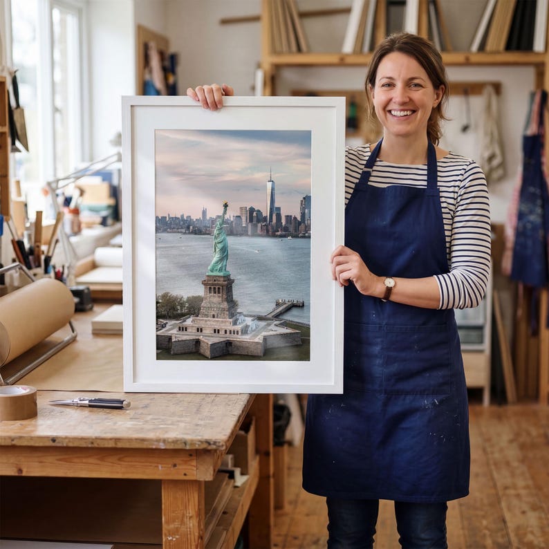 May include: A framed vertical photograph of the Statue of Liberty standing on Liberty Island with the New York City skyline and One World Trade Center visible in the background under a soft, light-filled sky during the daytime.