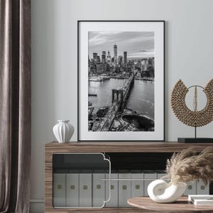 a framed black and white photograph of the Brooklyn Bridge, which is a prominent landmark in New York City. The photograph is displayed on a wall above a wooden cabinet or shelf, which has several books arranged on it. The overall setting appears to be a well-organized and aesthetically pleasing interior space.