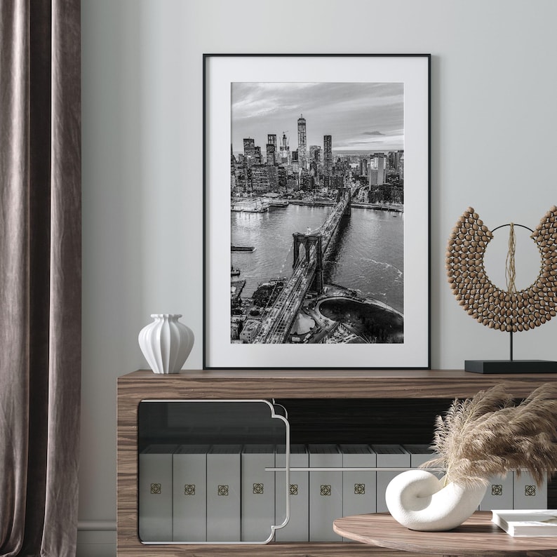 a framed black and white photograph of the Brooklyn Bridge, which is a prominent landmark in New York City. The photograph is displayed on a wall above a wooden cabinet or shelf, which has several books arranged on it. The overall setting appears to be a well-organized and aesthetically pleasing interior space.