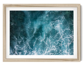 Op de afbeelding: Een ingelijste kunstdruk met een luchtfoto van diepblauw oceaanwater met witte schuimpatronen. Het kunstwerk is gevat in een lichtgekleurde houten lijst met een strakke, rechthoekige rand die de getextureerde afbeelding van het water omringt.