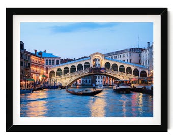 Può includere: Una stampa artistica incorniciata che raffigura il Ponte di Rialto a Venezia al crepuscolo. Il ponte in pietra ad arco attraversa il Canal Grande, illuminato da luci calde, con una gondola e barche sull'acqua. Il tutto è in una cornice nera.