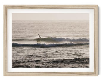 May include: A framed wall art print featuring a monochromatic photograph of a surfer riding a wave in the ocean. The scene captures the silhouette of the surfer against the light, with white foam breaking on the water and a light wood frame.