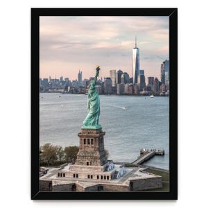 May include: A framed photographic print featuring the Statue of Liberty in a verdigris green patina standing on a stone pedestal, with the New York City skyline and One World Trade Center visible in the background under a soft sunset sky.