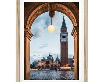 May include: A framed wall art print featuring a view of St. Mark's Basilica and its bell tower in Venice, Italy, seen through a stone archway with a hanging globe light fixture. The light wood frame surrounds the vertical architectural photograph.