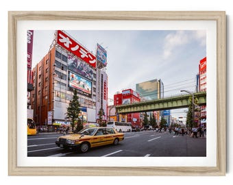 Op de afbeelding: Een ingelijste foto van een drukke straat in Tokio met een gele taxi op de voorgrond, gebouwen van meerdere verdiepingen met grote rode en witte Japanse uithangborden, een groen verhoogd spoorwegviaduct en voetgangers op de weg.