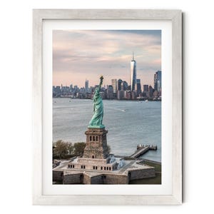 May include: A framed wall art print featuring the Statue of Liberty standing on its star-shaped pedestal in New York Harbor, with the Manhattan skyline visible in the background under a soft, pale sky at dusk, enclosed in a light-colored wooden frame.