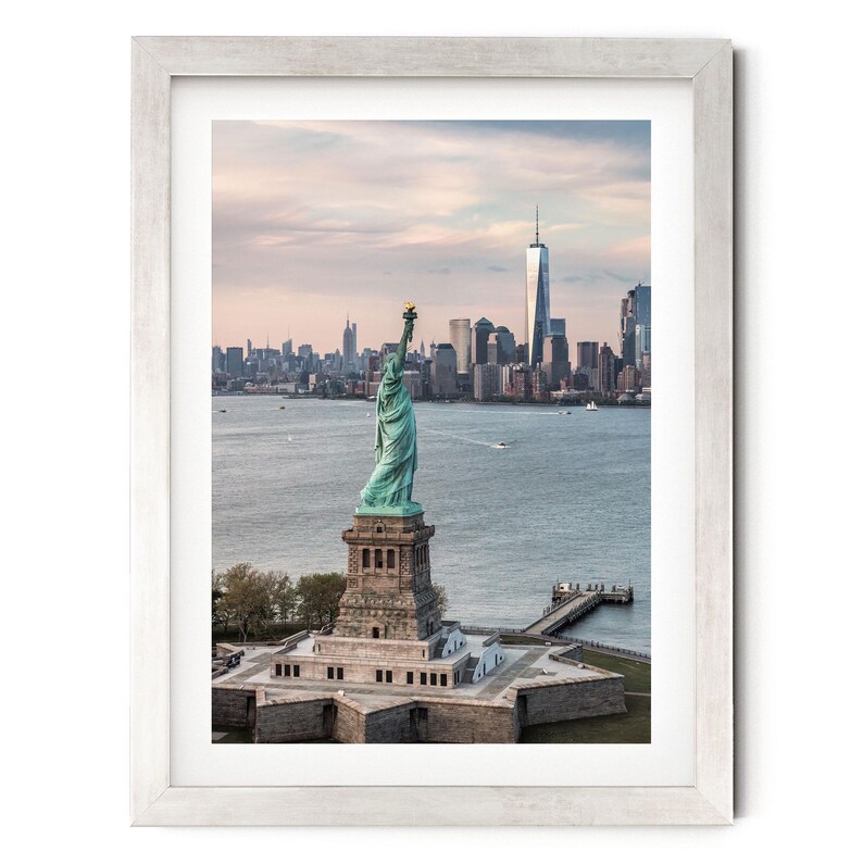 May include: A framed wall art print featuring the Statue of Liberty standing on its star-shaped pedestal in New York Harbor, with the Manhattan skyline visible in the background under a soft, pale sky at dusk, enclosed in a light-colored wooden frame.