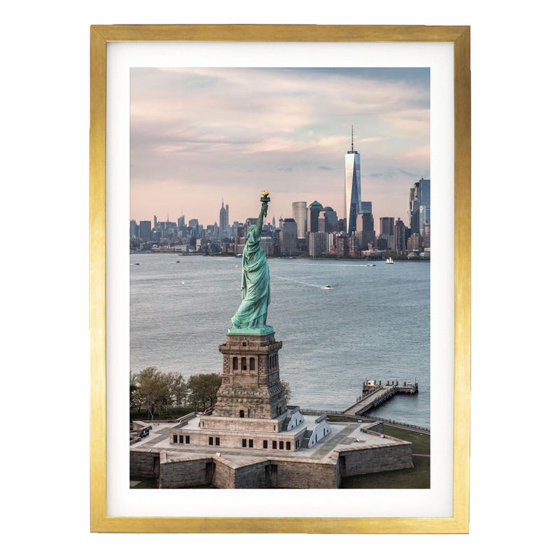 May include: A framed wall art print featuring the Statue of Liberty standing on its star-shaped pedestal in New York Harbor, with the Manhattan skyline and One World Trade Center visible in the background under a soft, pale sunset sky.
