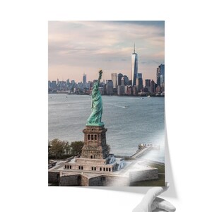 May include: A photographic print featuring the Statue of Liberty standing on its pedestal in New York Harbor, with the Manhattan skyline and One World Trade Center visible in the background under a soft, overcast sky during the daytime.